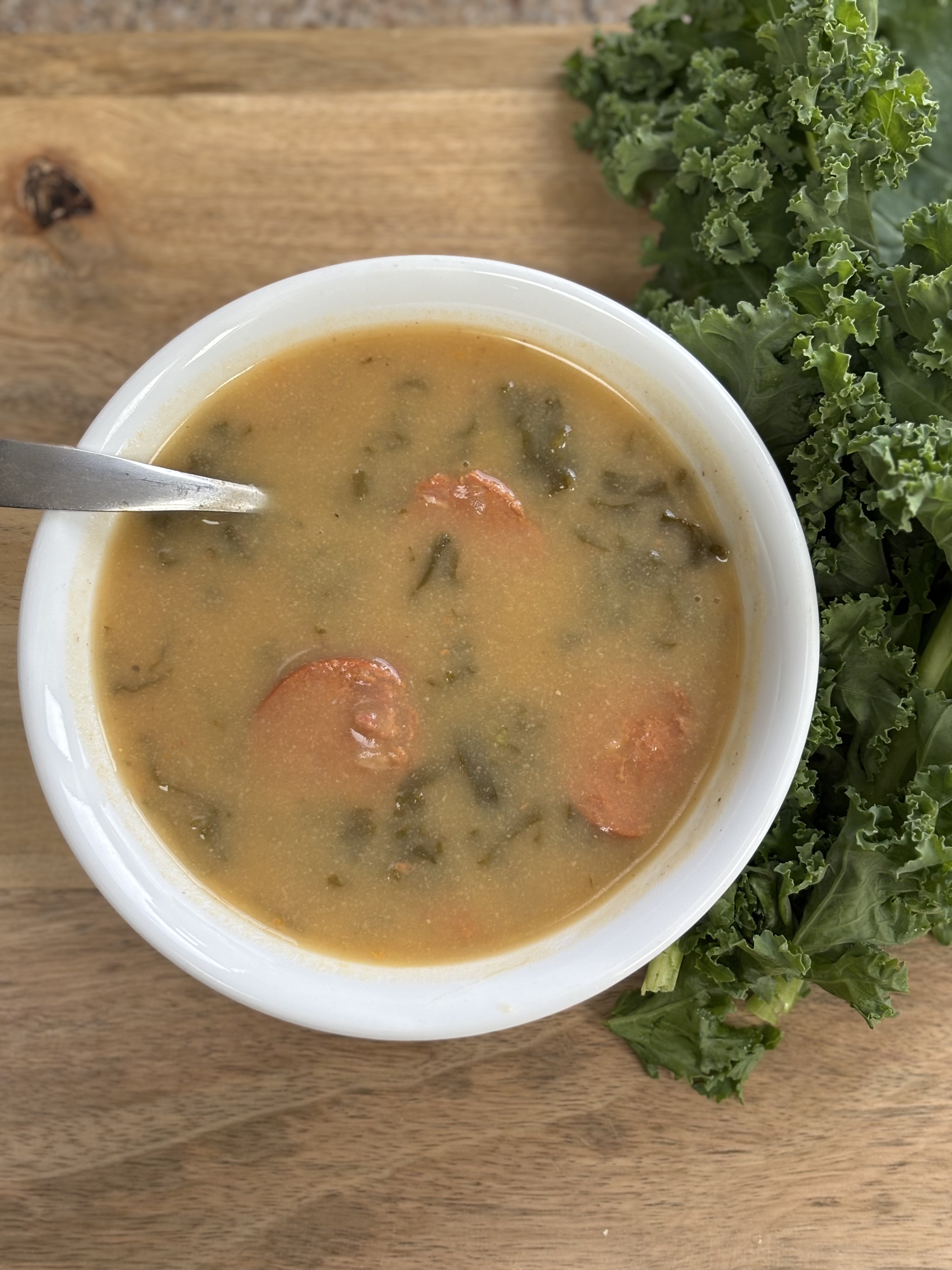 Caldo Verde Portuguese kale soup with sliced chorizo in a white bowl alongside fresh kale leaves on a wooden board
